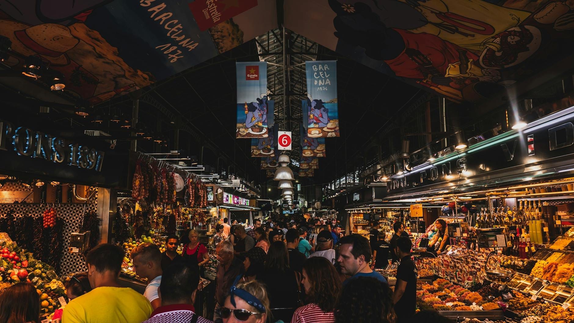 Busy indoor food market in Barcelona with crowds of people and colorful produce stalls under banners.
