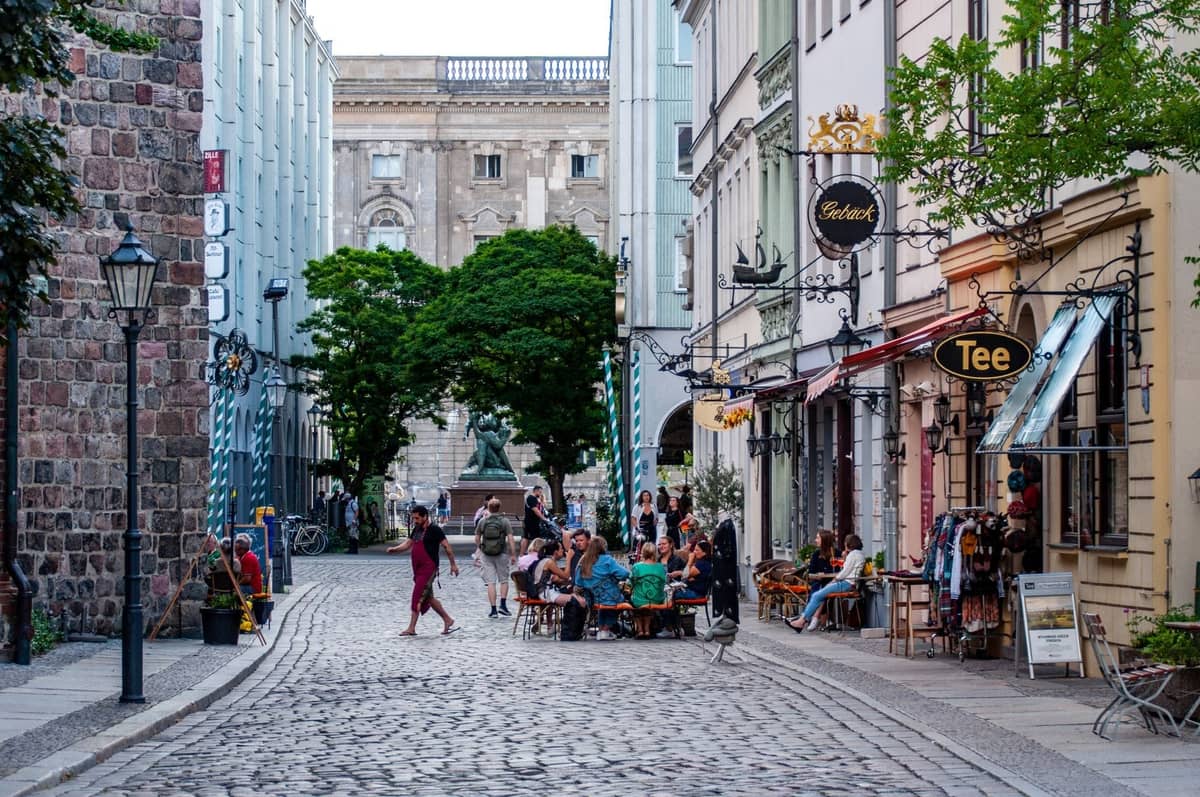 Kopfsteinpflasterstraße in Berlin, gesäumt von Cafés und kleinen Geschäften, Menschen beim Essen im Freien und ein historisches Gebäude im Hintergrund.