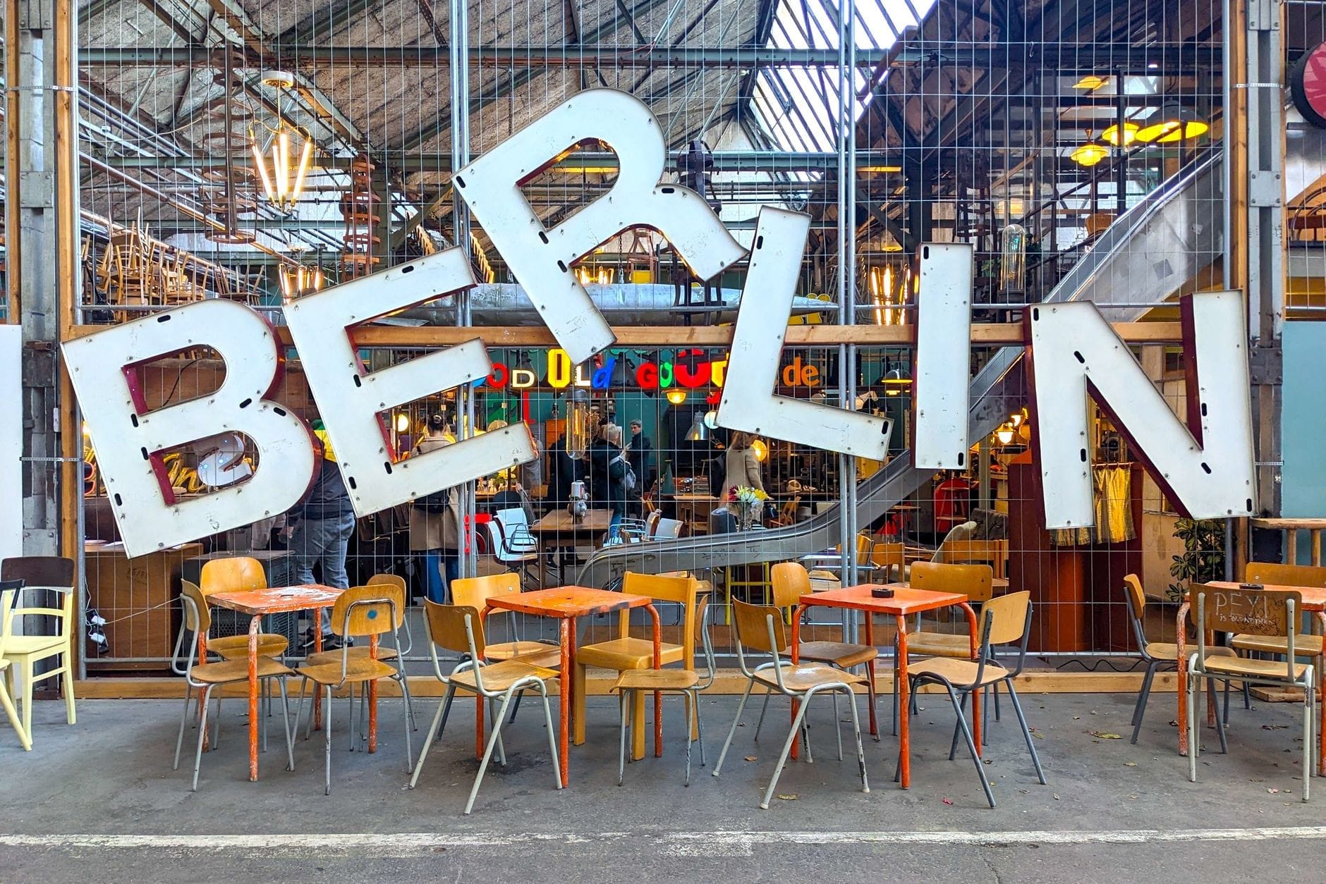 Mesas coloridas ao ar livre em um animado food hall de Berlim, com uma grande placa divertida de BERLIN acima das mesas e frequentadores dentro atrás de uma grade de arame.
