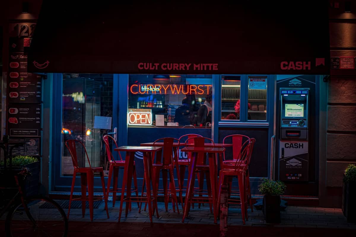 Nächtliche Straßenansicht eines Berliner Currywurst-Stands namens Cult Curry Mitte, mit Leuchtreklamen und roten Stühlen draußen.