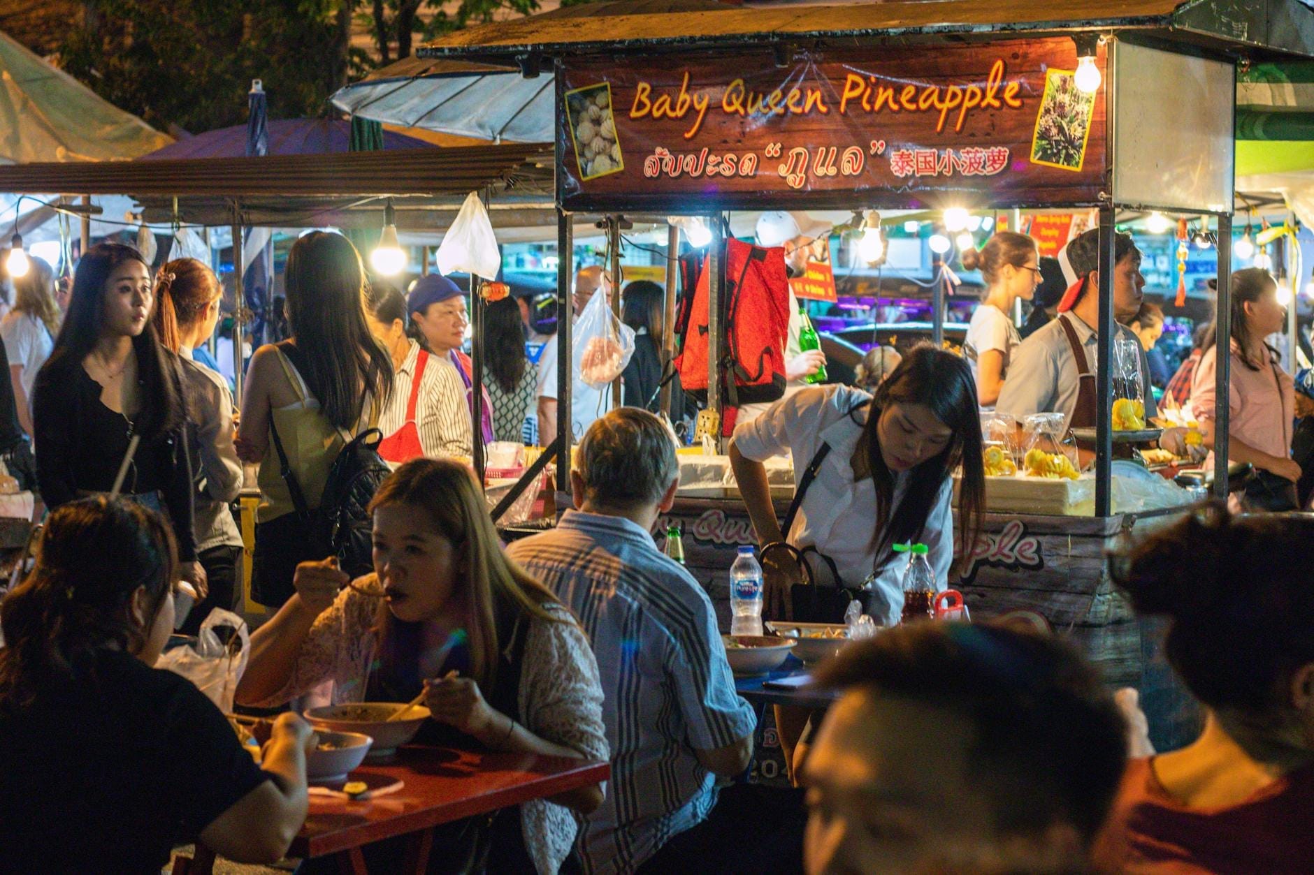Animada escena del mercado nocturno de Chiang Mai con personas comiendo en mesas y comprando en puestos de comida iluminados.