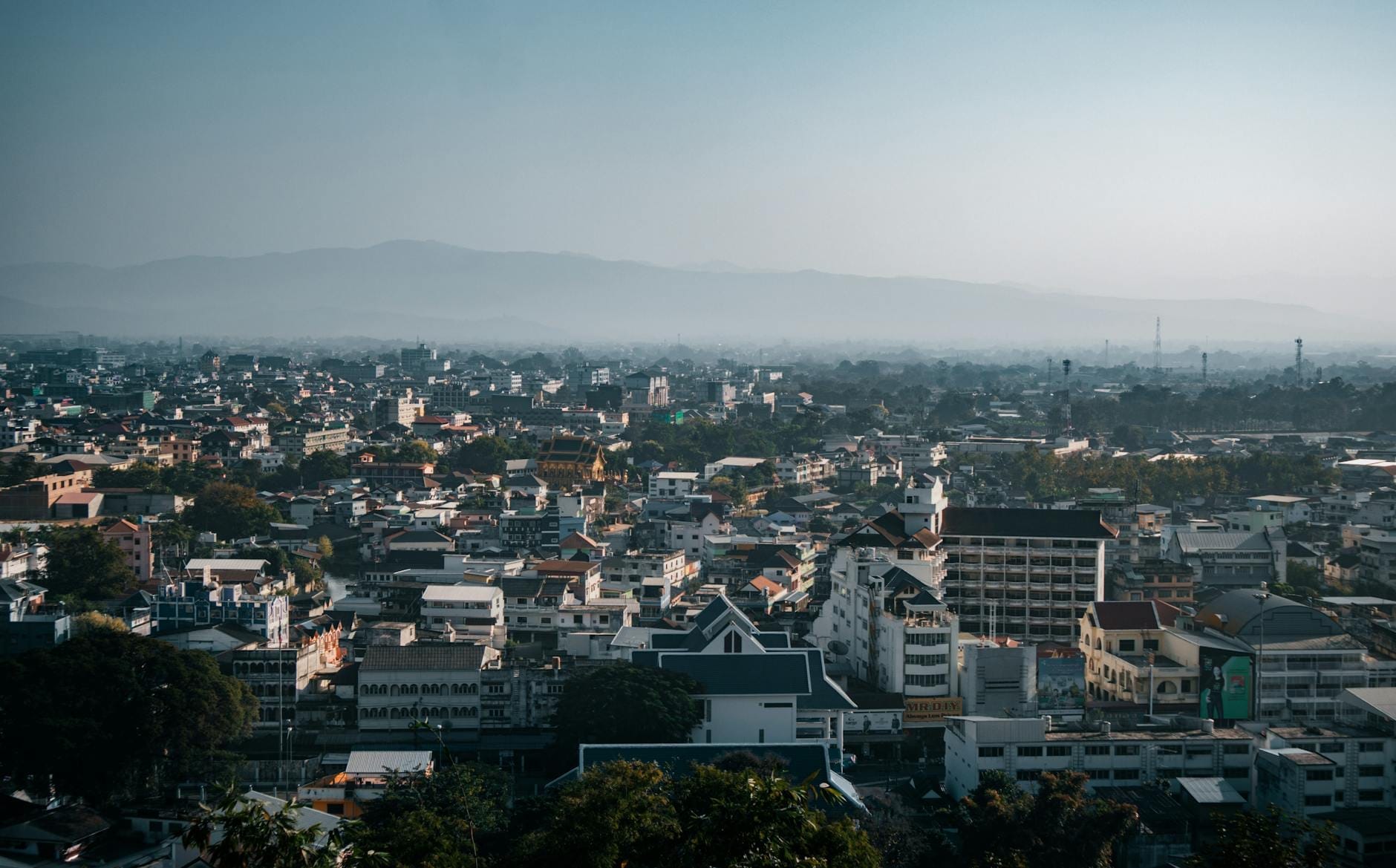 Vista aérea amplia de la ciudad de Chiang Mai que muestra el trazado urbano con montañas distantes bajo un cielo neblinoso.