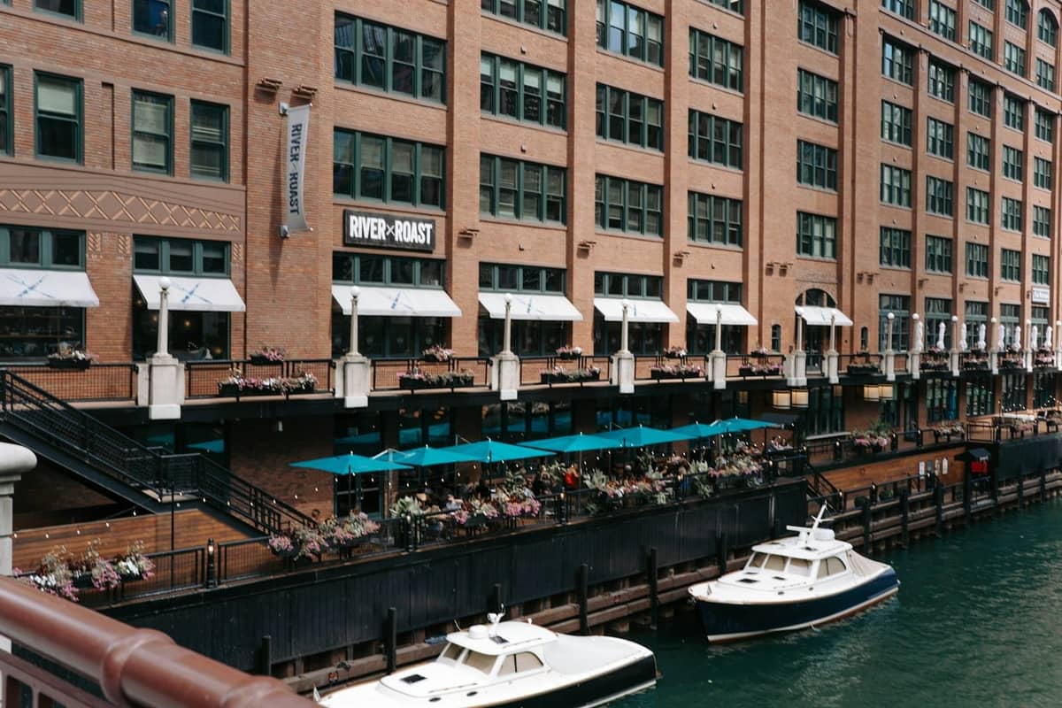 Chicago Riverwalk con posti a sedere all'aperto di un ristorante affollato di persone accanto all'acqua, barche ormeggiate nelle vicinanze e gli edifici della città sullo sfondo.