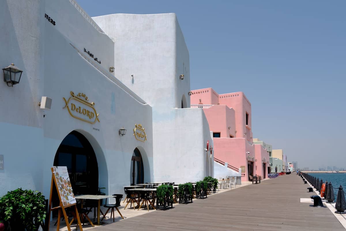 Bunte Restaurants und Cafés entlang der Promenade des Katara Cultural Village in Doha, mit Tischen im Freien und Blick auf das Meer unter strahlend blauem Himmel.