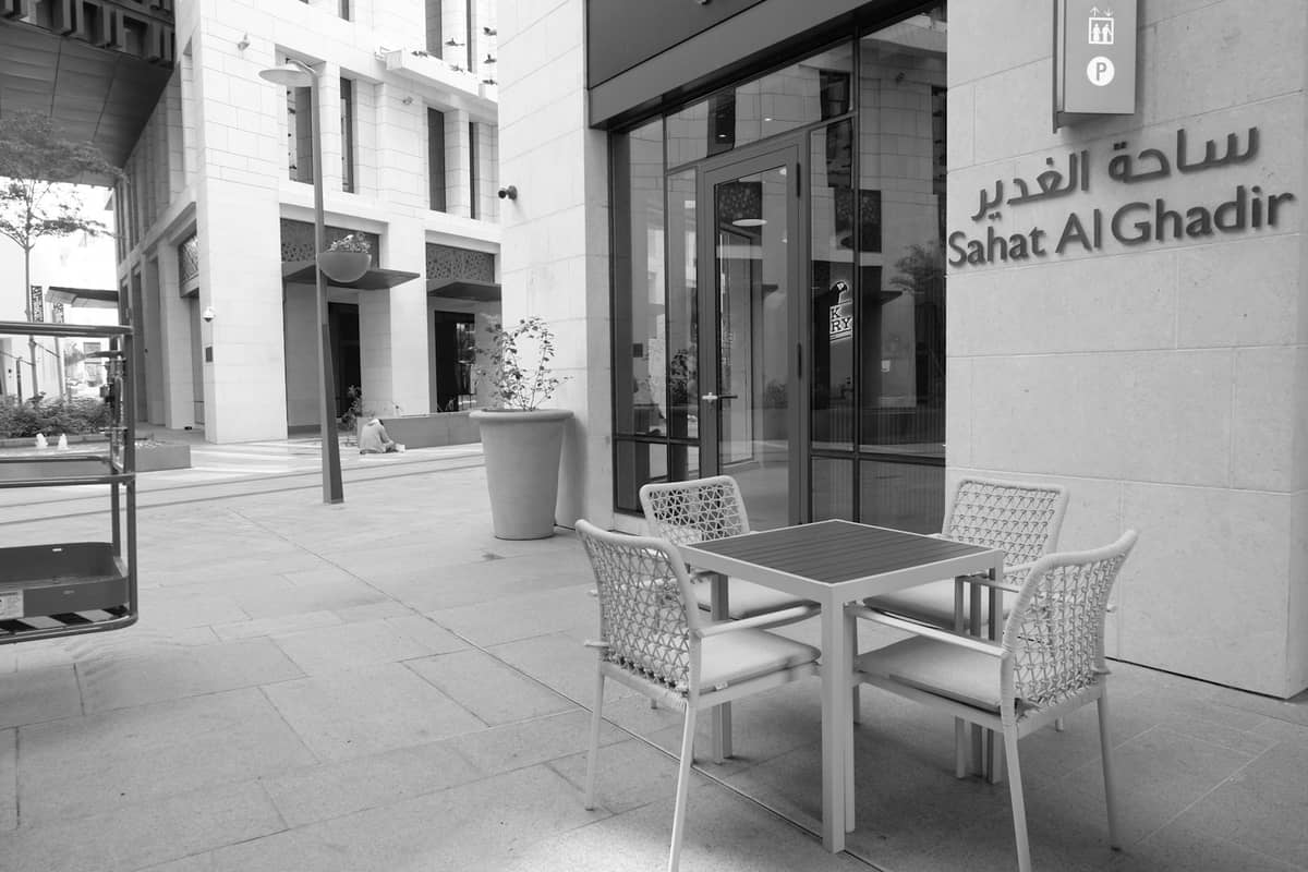 Monochromer Blick auf ein modernes Außencafé in Msheireb Downtown Doha, mit schlichter Architektur und arabisch-englischem Schild 'Sahat Al Ghadir'.