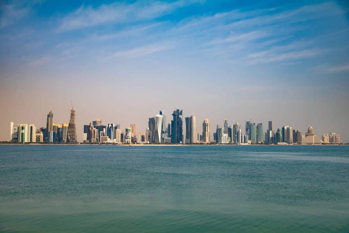 Panoramablick auf die Skyline von West Bay in Doha mit Hochhäusern entlang der Corniche, klarem blauen Himmel und ruhigem Wasser im Vordergrund.