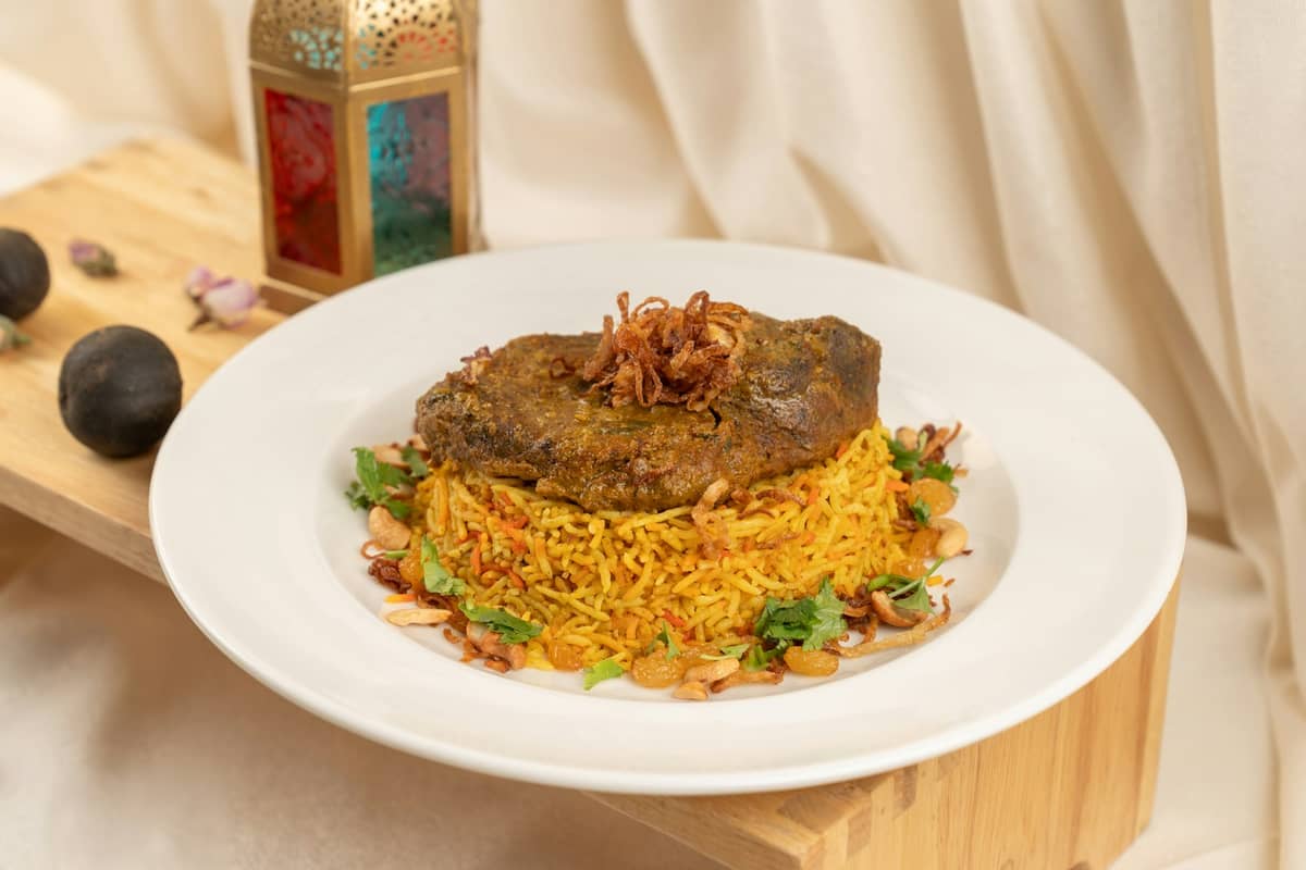 Ein Teller traditionell gewürzten Reises mit Fleisch, garniert mit Nüssen und Kräutern, neben getrockneten Limetten und einer dekorativen Laterne.
