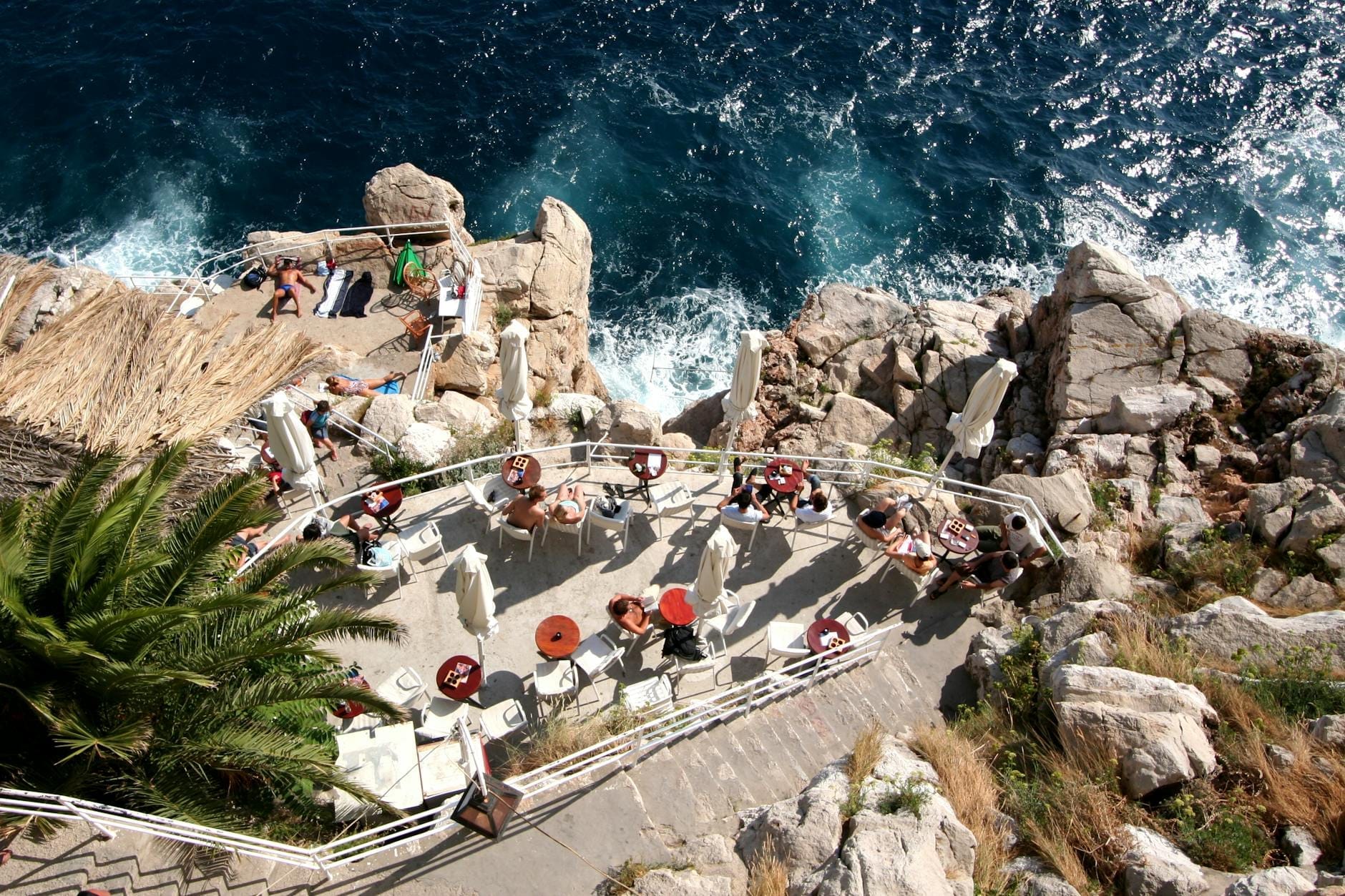 Vista aérea de la terraza de un restaurante junto al mar sobre acantilados rocosos en Dubrovnik, con personas cenando y olas del mar azul abajo.