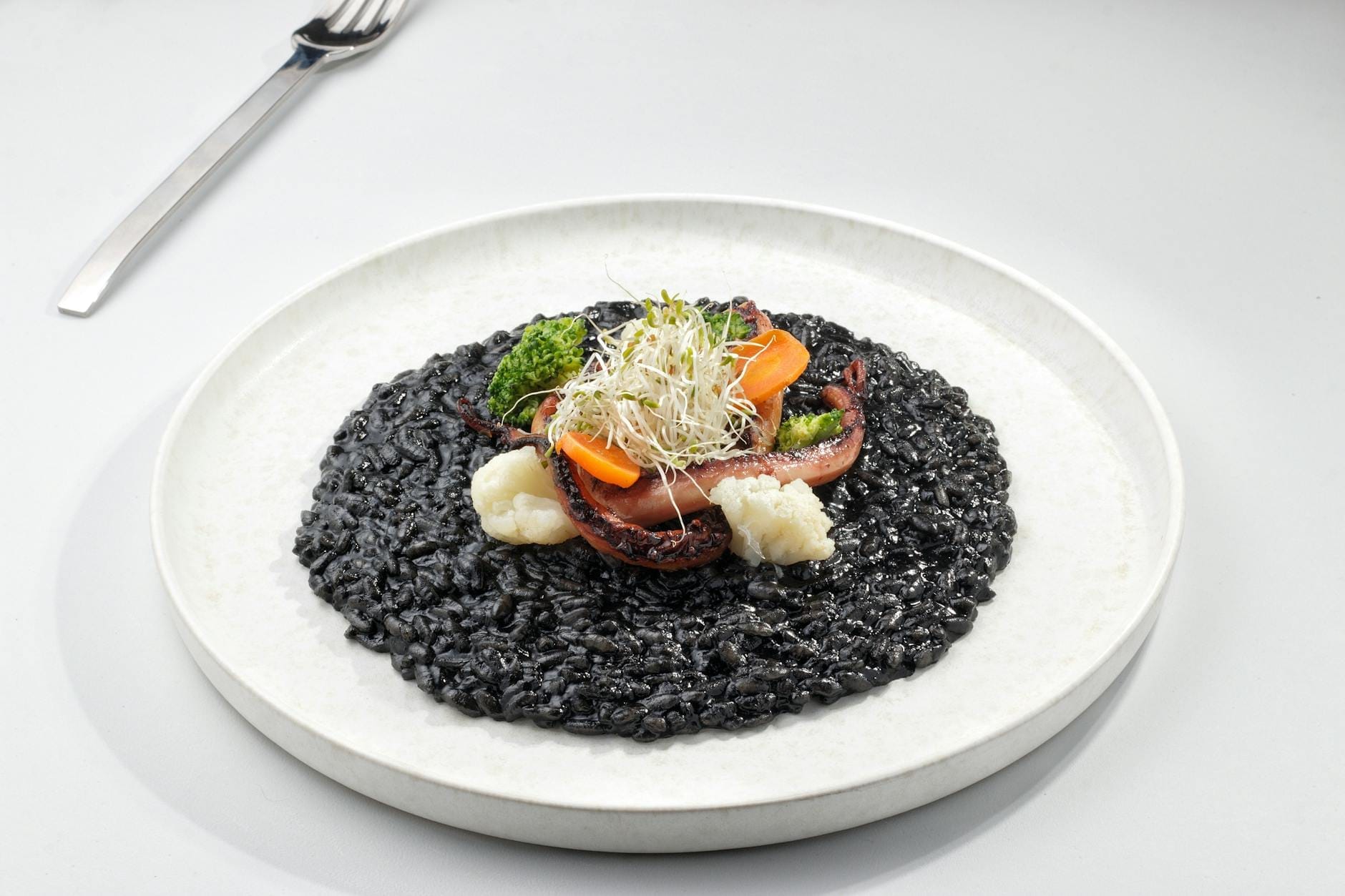 Un plato de risotto negro garnecido con verduras y brotes, presentado en una mesa blanca con un tenedor cerca.