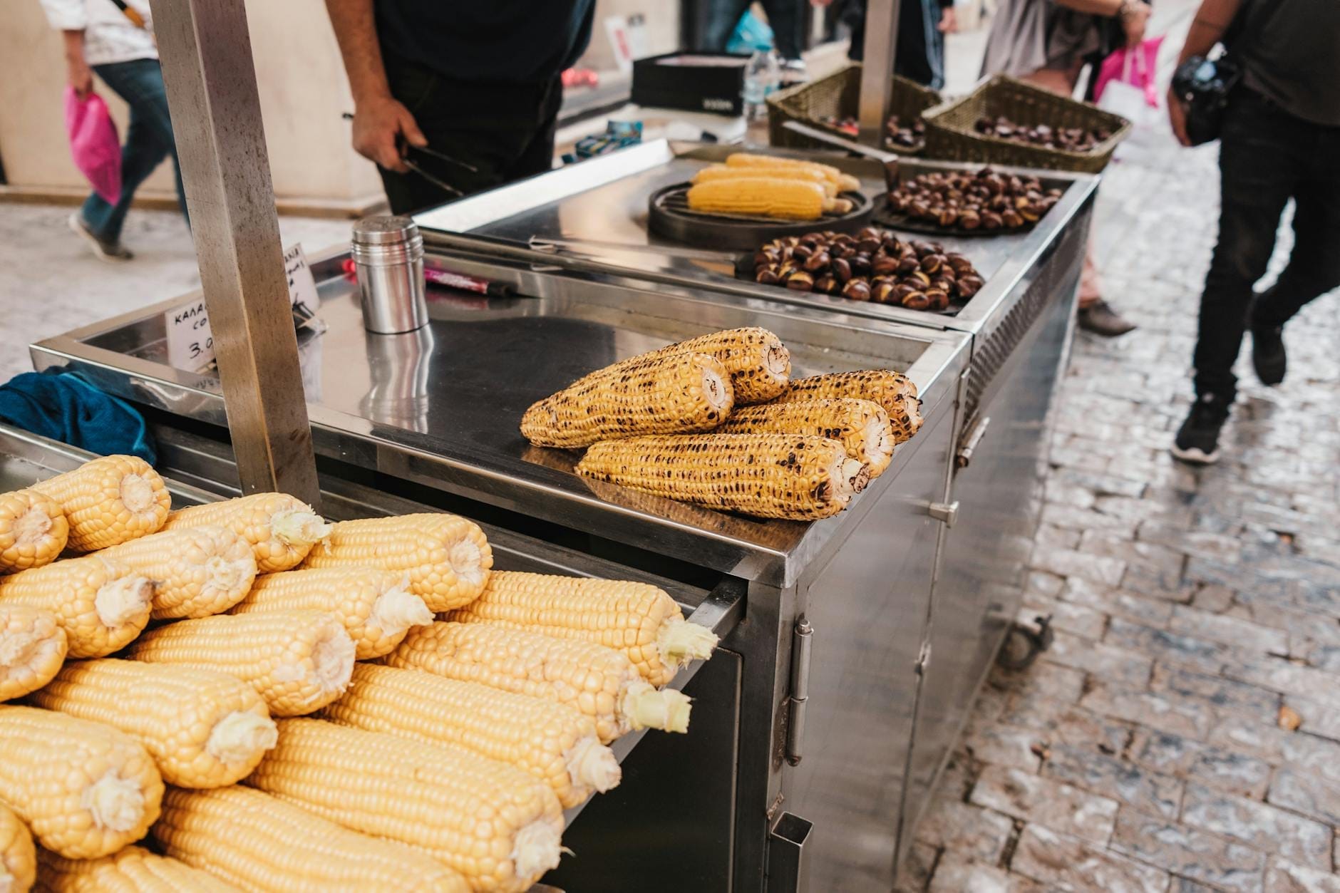 Puesto de comida callejera con maíz a la parrilla y castañas asadas en una calle adoquinada, con personas caminando cerca