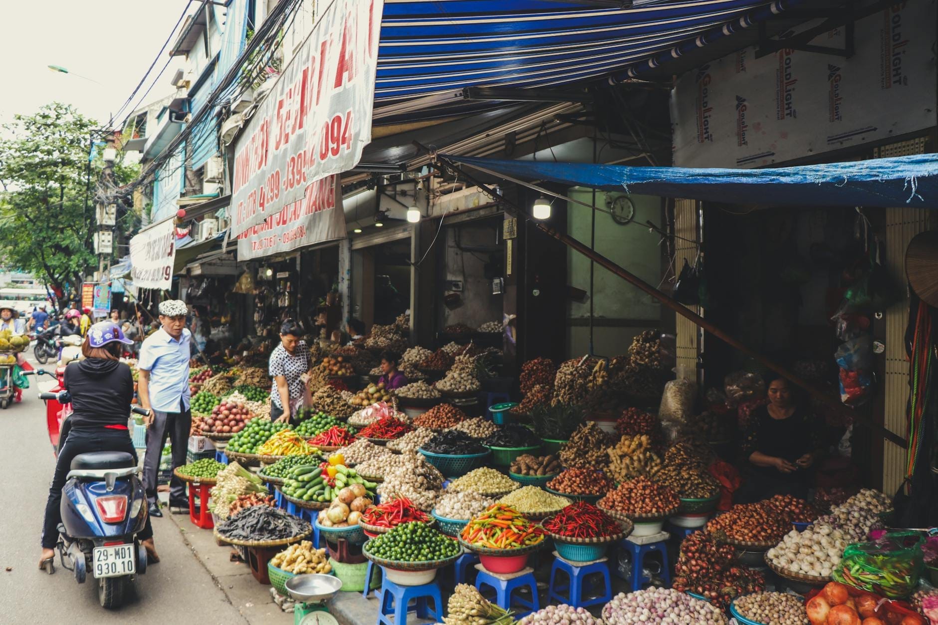 Bustling street market in Hanoi with colorful produce and vendors on small stools, shoppers and scooters passing by fresh fruit and vegetables.