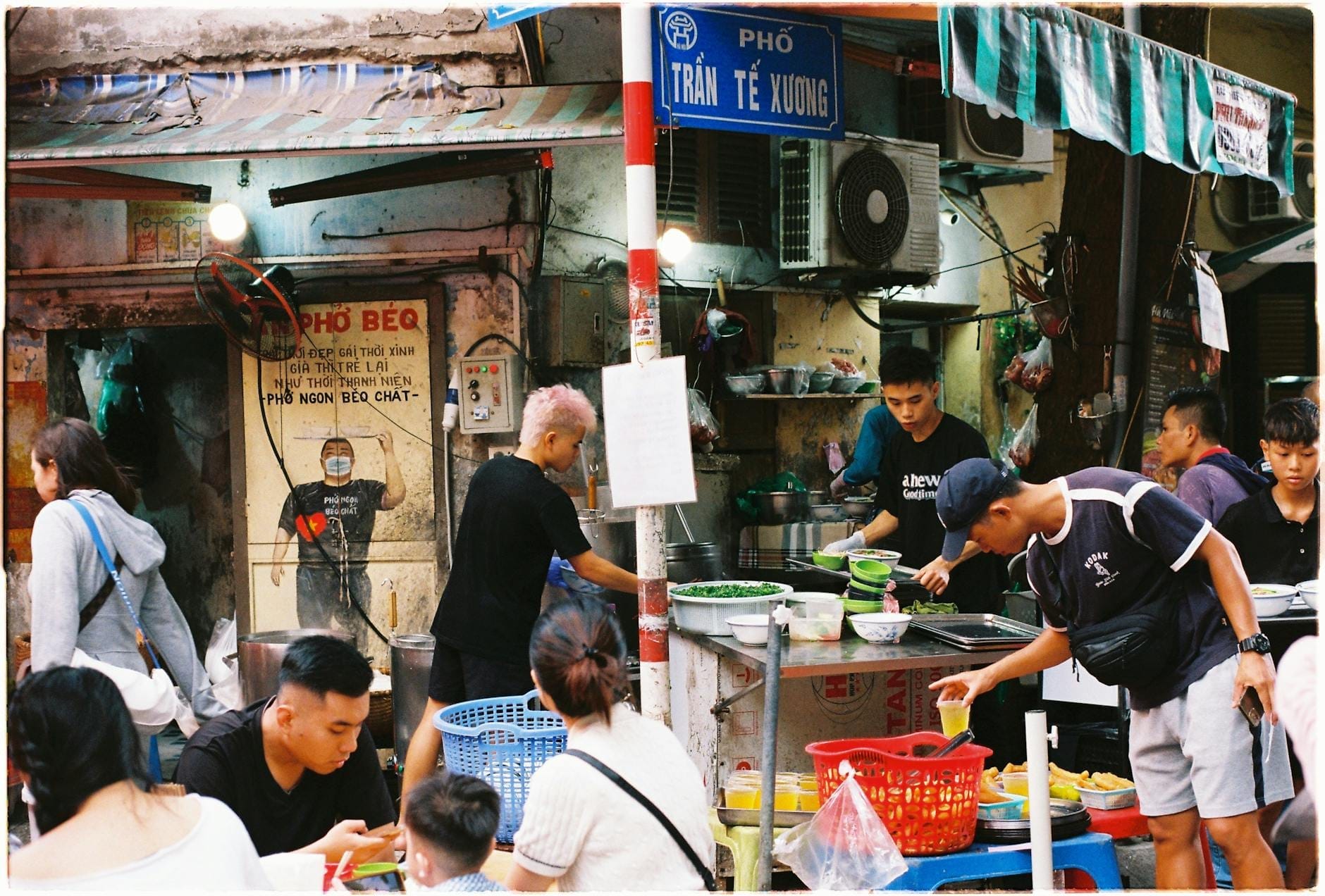 Busy Hanoi street food stall with locals eating pho at blue tables, kitchen action, and prominent street signs in Vietnamese.