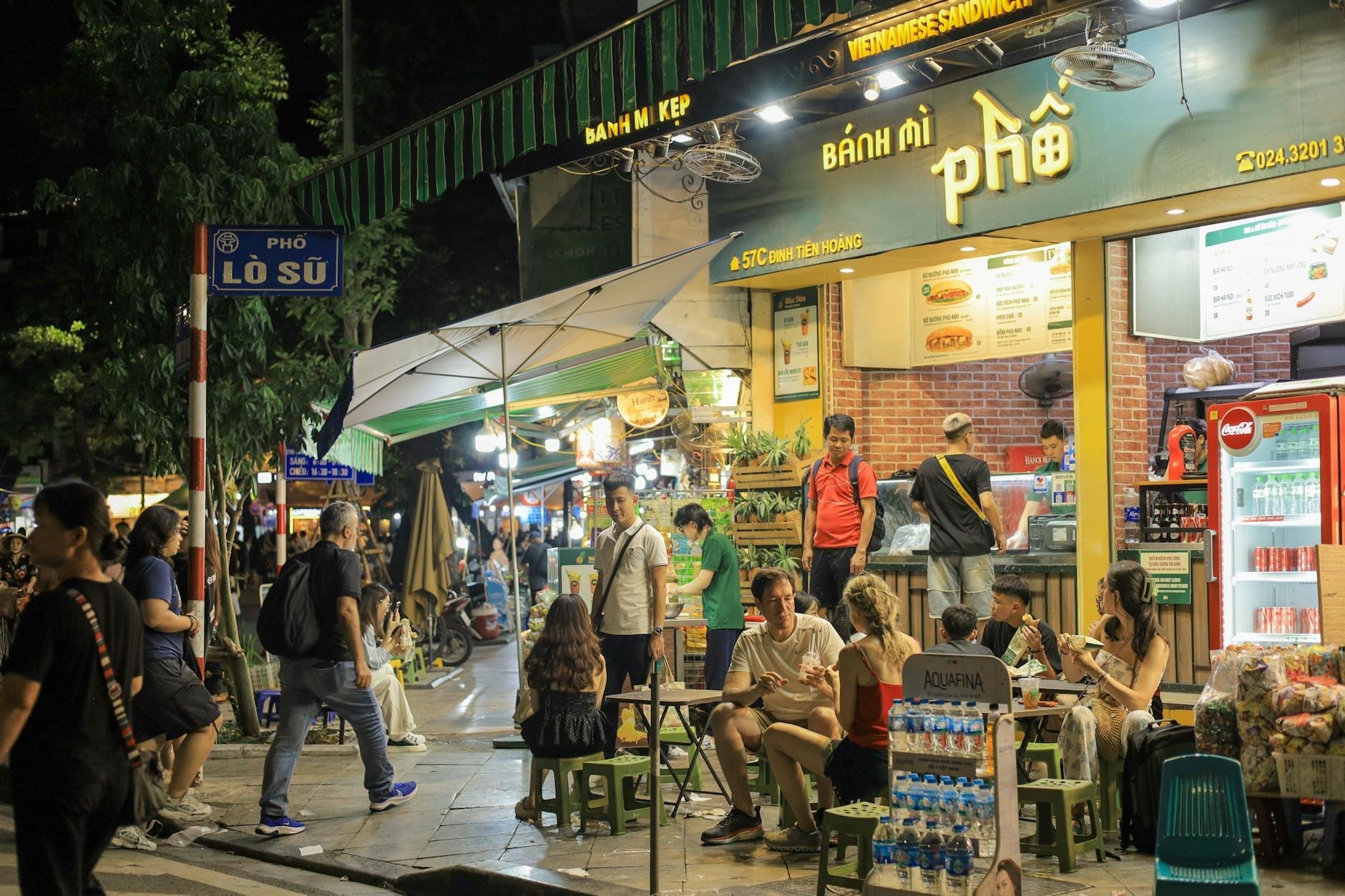 A lively Hanoi street scene at night with locals and tourists eating at outdoor tables in front of street food stalls and restaurants.