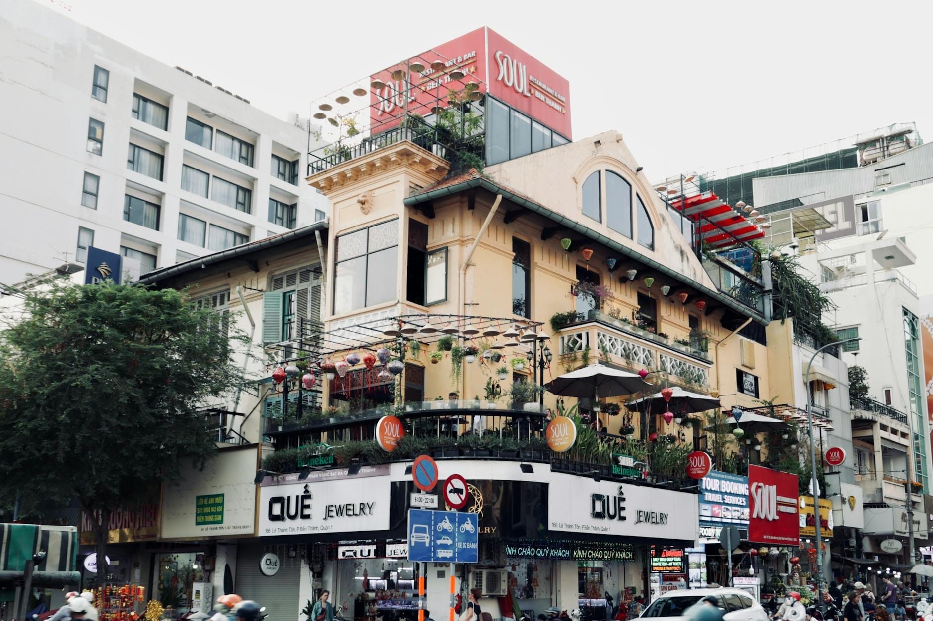 Animada esquina de una calle de Saigón con un edificio de varios pisos que alberga restaurantes, señales de tráfico, vegetación y personas caminando en el centro de Ho Chi Minh City.