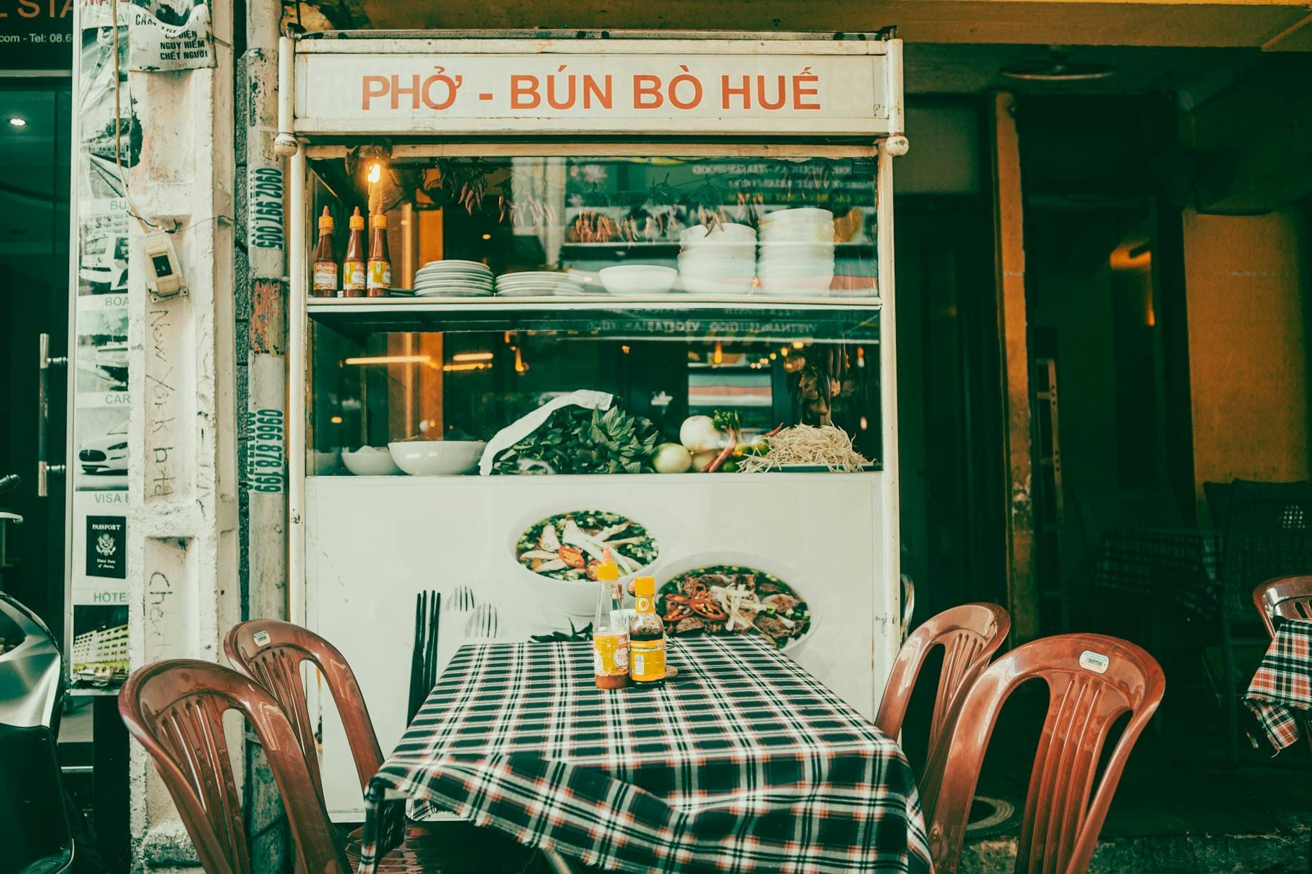 Un puesto clásico de comida callejera vietnamita con un letrero que dice 'Phở - Bún Bò Huế', con sillas de plástico y una mesa a cuadros en el exterior.