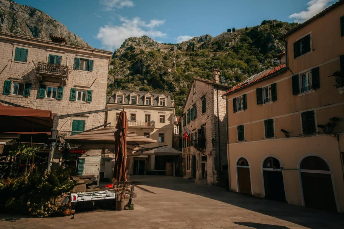 An inviting square in Kotor Old Town with outdoor tables and umbrellas, surrounded by medieval stone buildings and mountains in the background.