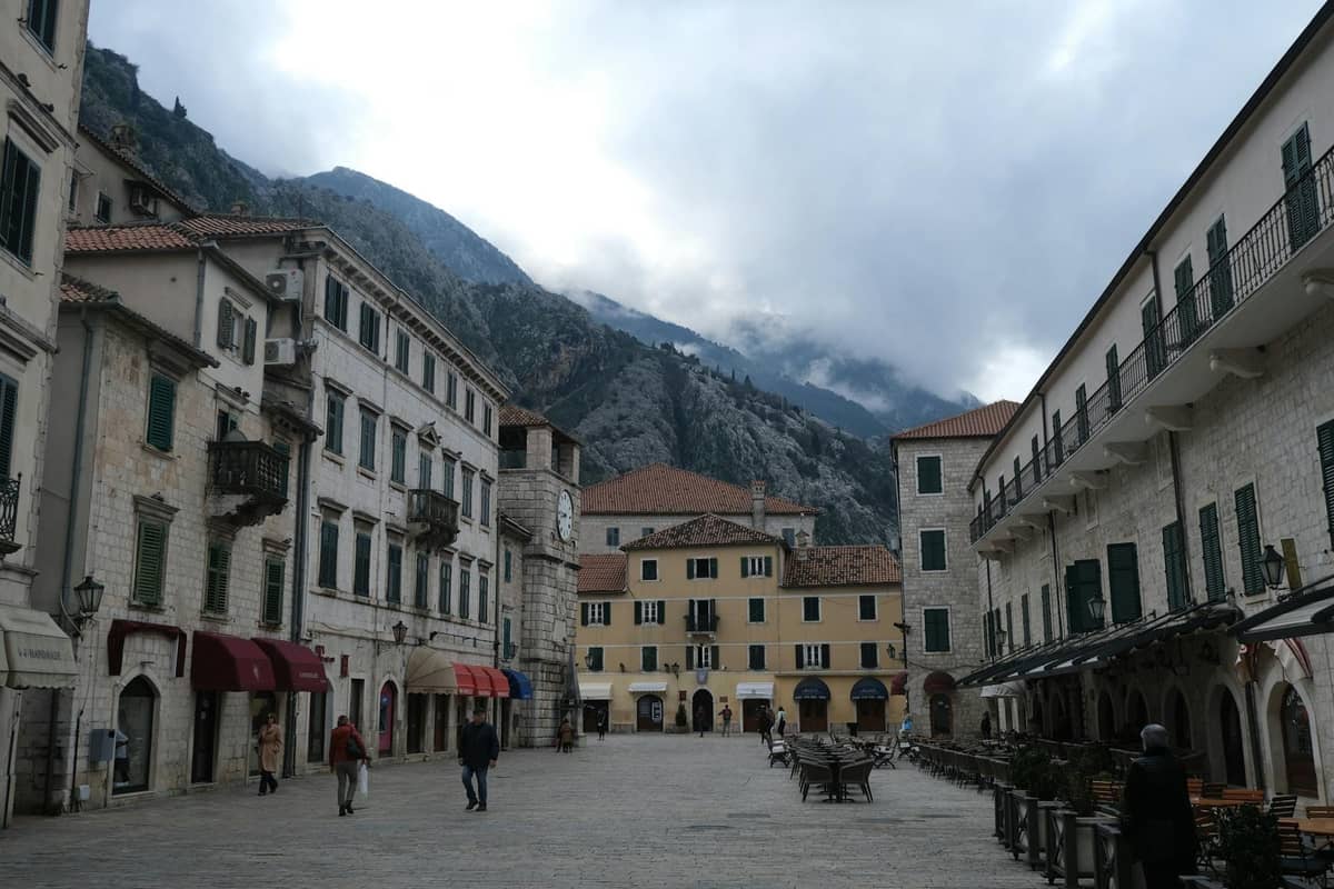 Wide stone plaza in Kotor old town with outdoor cafe seating, historic buildings, mountains in background, and a few people walking.