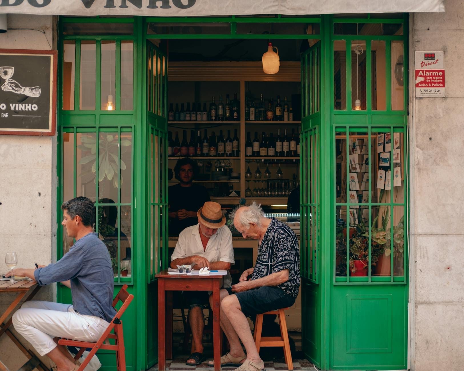 Charmant petit restaurant de Lisbonne avec des portes vertes, des étagères de bouteilles de vin et des personnes assises à des tables à l'intérieur comme à l'extérieur.