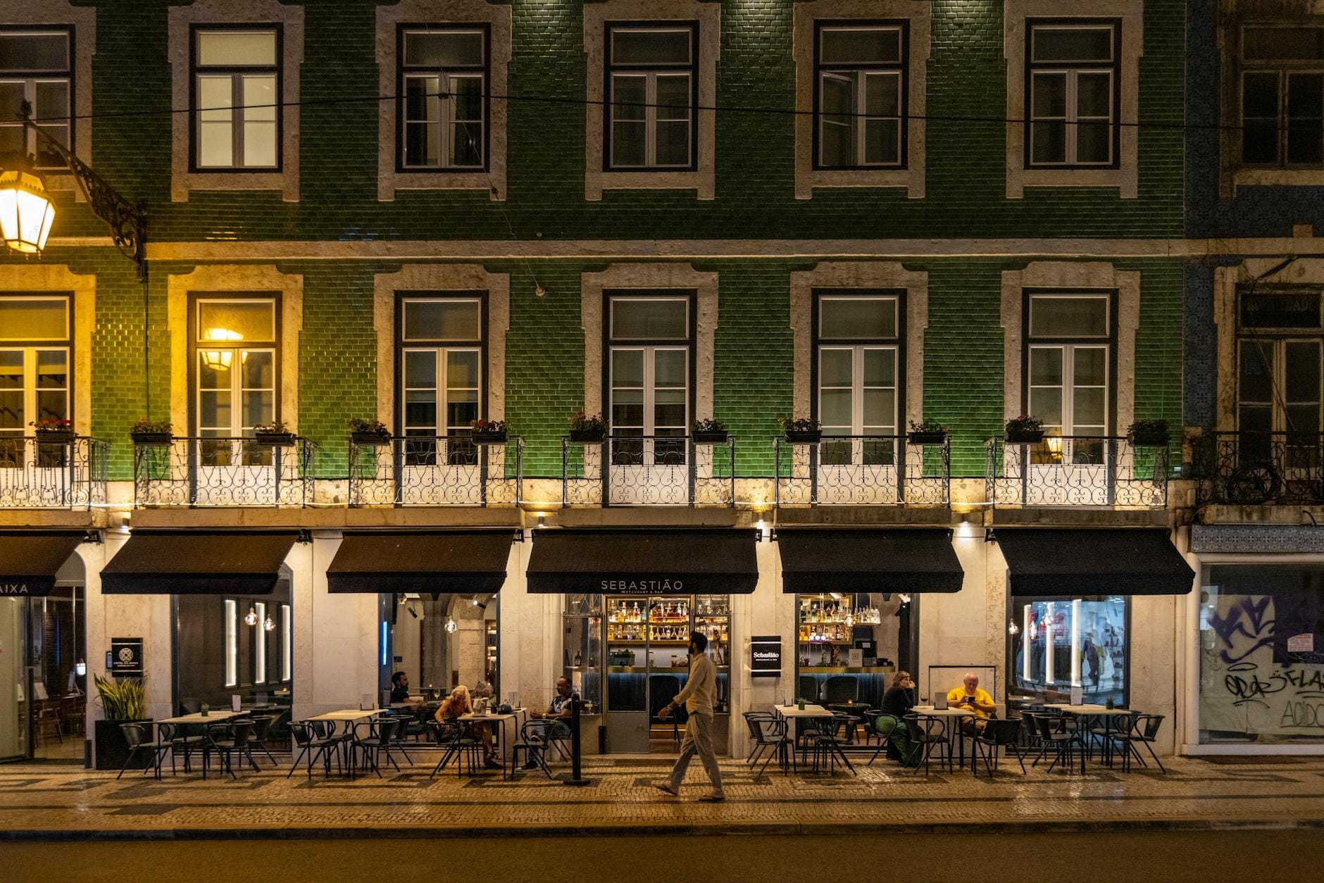 Vue nocturne d'un élégant restaurant de Lisbonne avec terrasse extérieure, façade en carreaux verts et des gens dînant dans une rue animée de la ville.