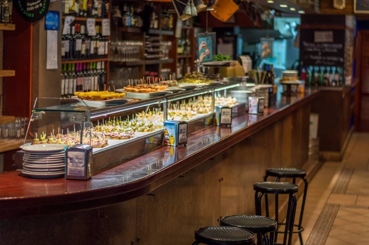 Larga barra dentro de un restaurante español de tapas con varios pintxos y tapas en exposición, taburetes a lo largo del mostrador y botellas de vino detrás.