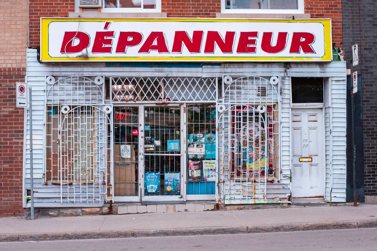 Straßenansicht eines Dépanneurs in Montreal mit einem großen rot-gelben Schild, vergitterten Fenstern und Plakaten im Fenster.