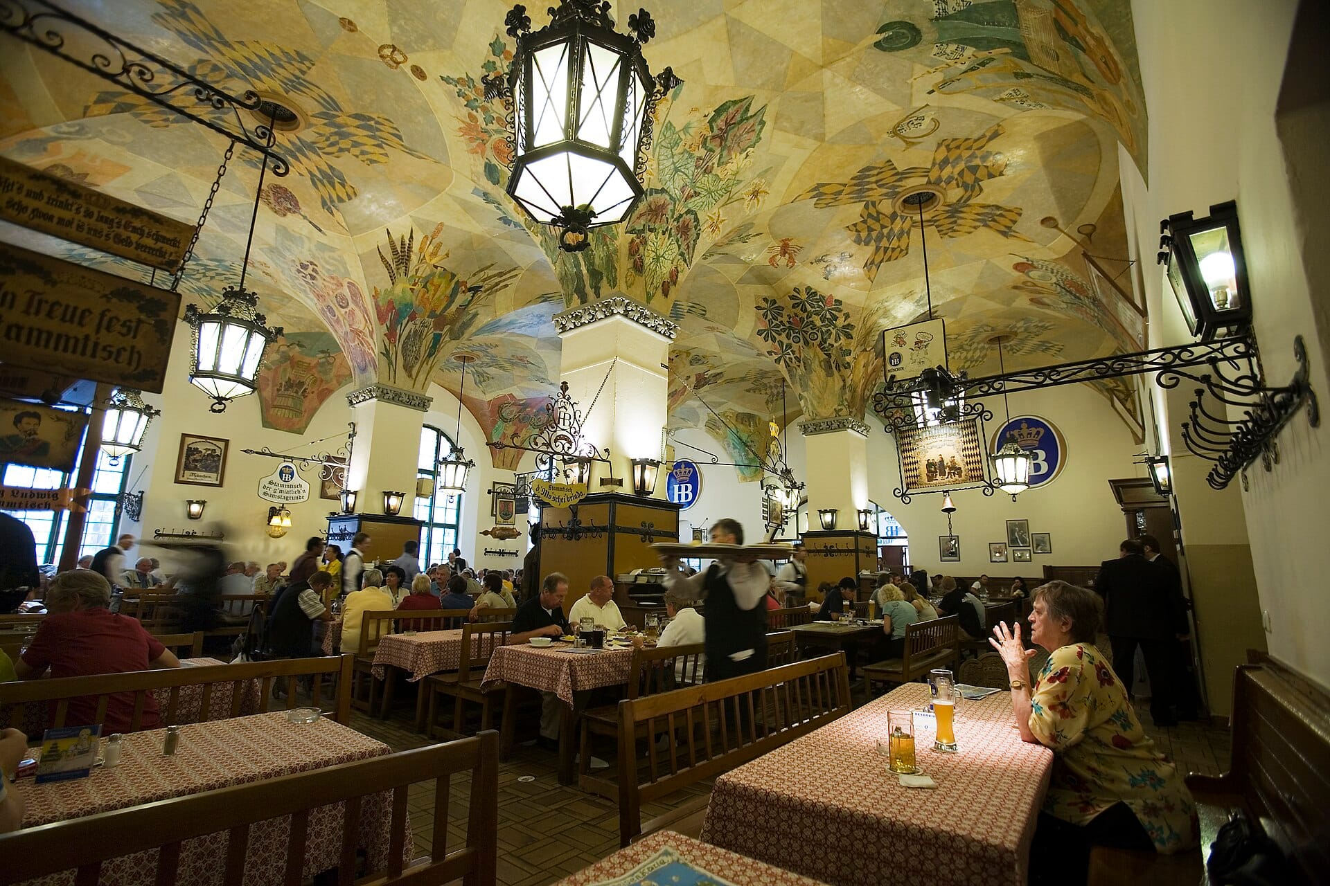 บรรยากาศคึกคักภายในห้องโถงเบียร์ Hofbräuhaus ในมิวนิก พร้อมเพดานโค้งทาสีขนาดใหญ่ ม้านั่งไม้ และผู้คนที่เพลิดเพลินกับเครื่องดื่มที่โต๊ะ