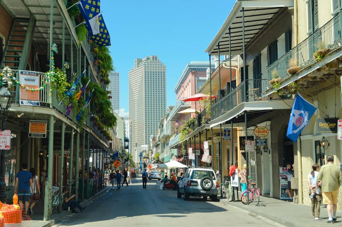 Bustling street scene in New Orleans with classic wrought-iron balconies, flags, restaurants, and people walking under a blue sky.
