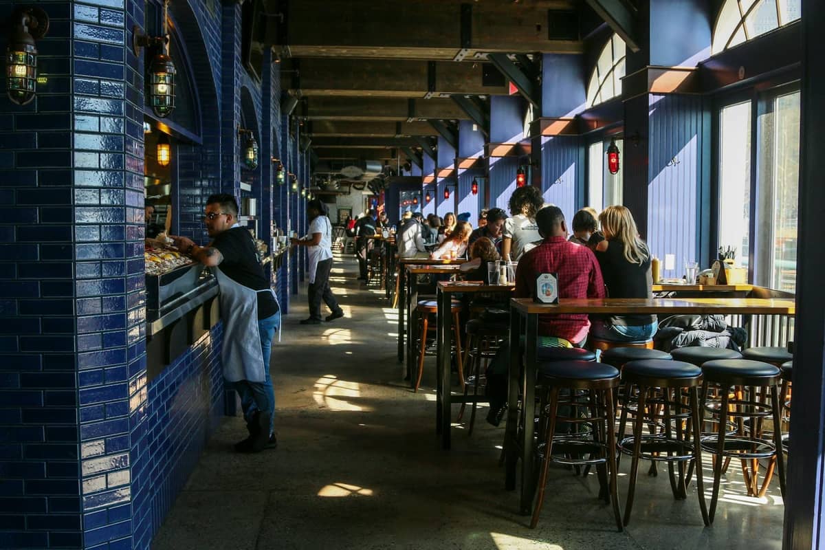 A bustling, sunlit restaurant with blue tiled walls, diners enjoying meals, and staff serving food, evoking the vibrant dining culture of New Orleans.