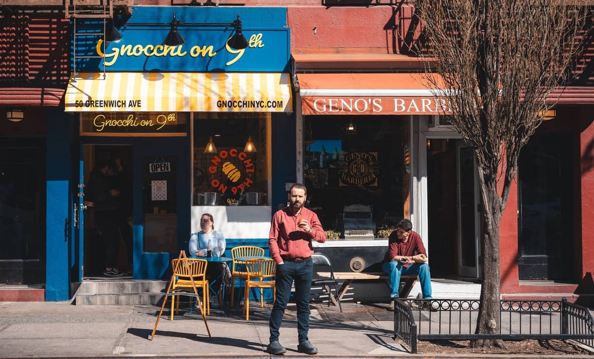 Straßenansicht zweier kleiner Restaurants in New York mit Außenbestuhlung, blau-gelbem Sonnendach, Fußgängern und städtischer Atmosphäre.