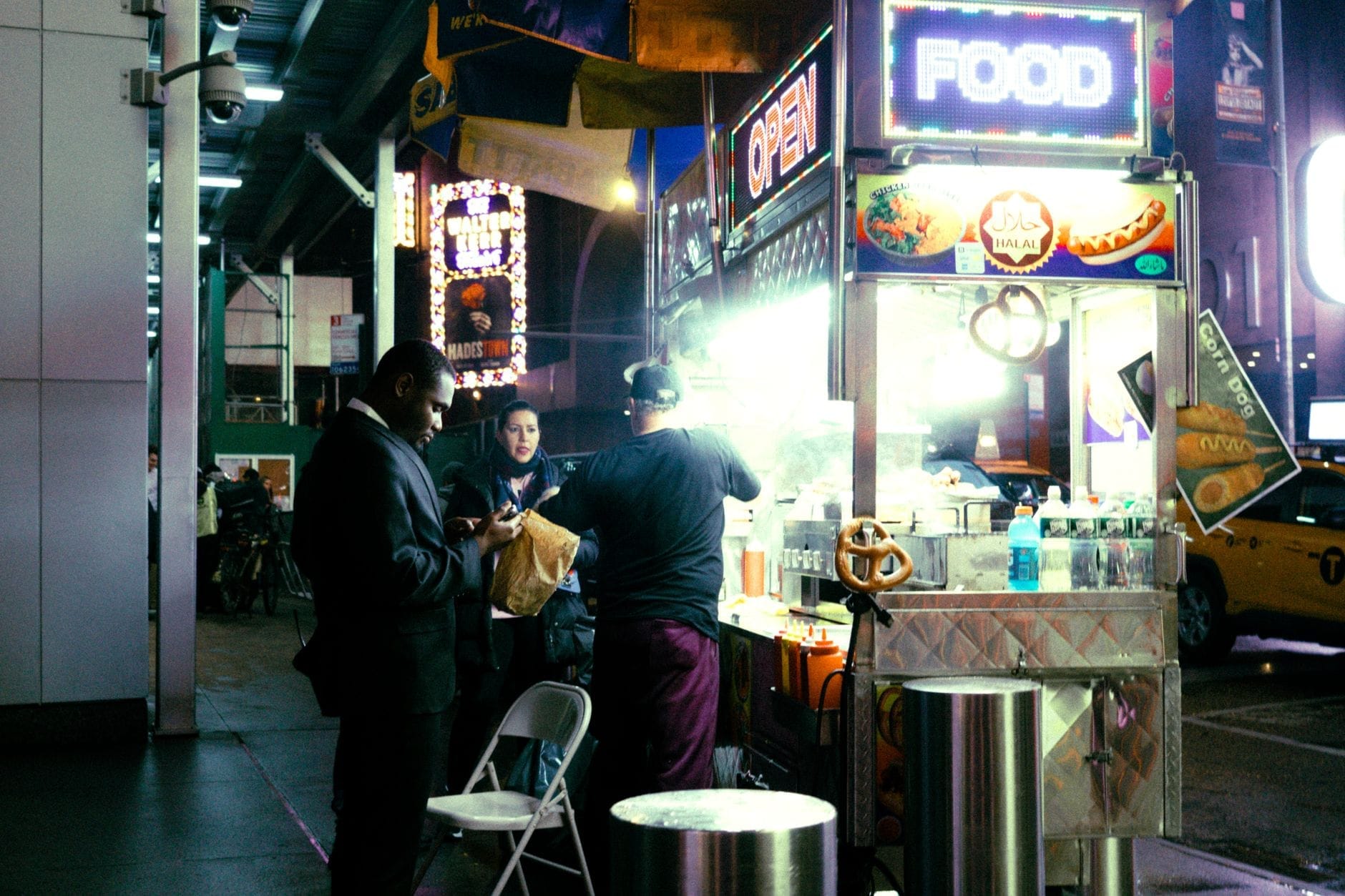 Uma barraca de comida de rua em Nova York à noite, com pessoas pedindo comida sob letreiros de neon brilhantes, capturando a diversidade gastronômica e a cultura das ruas nova-iorquinas.