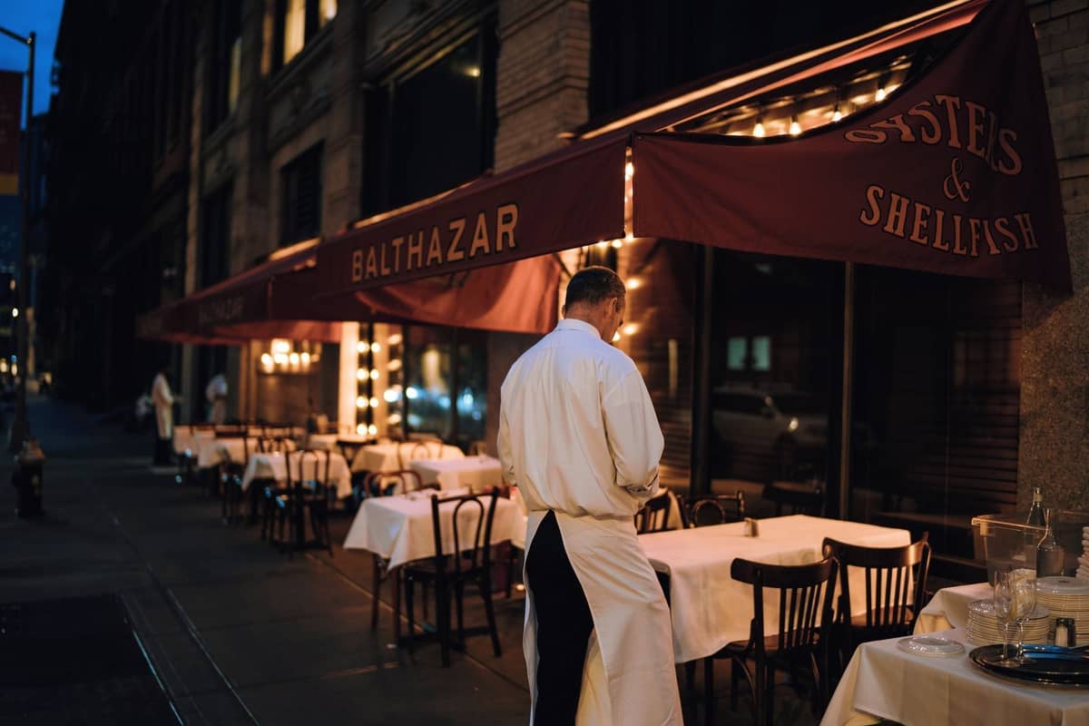 Kellner in weißer Schürze bereitet Außentische im Balthazar vor, einem bekannten Fine-Dining-Restaurant in New York City mit roten Markisen in der Abenddämmerung.