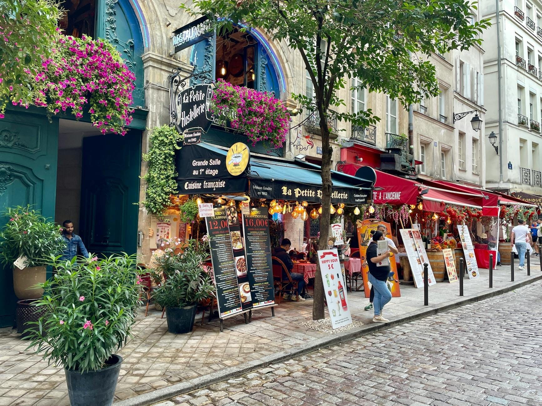 Belebte Pariser Straße gesäumt von Cafés und Restaurants, Speisekarten und Schilder draußen, Menschen beim Essen und Spazierengehen, Kopfsteinpflaster.