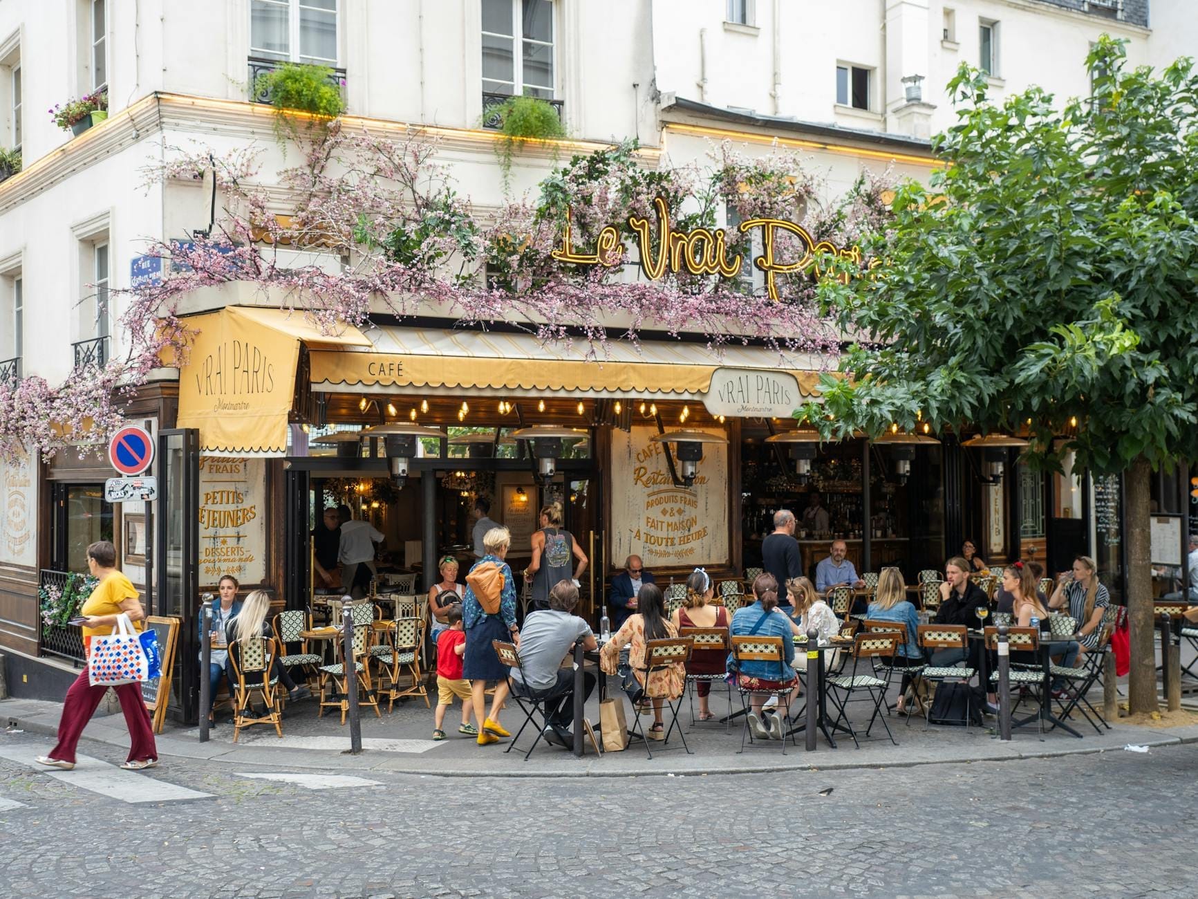 Un vivace caffè parigino con persone sedute ai tavoli all'aperto, decorazioni floreali sulla facciata e un'atmosfera di strada incantevole, perfetta per chi ama il cibo.
