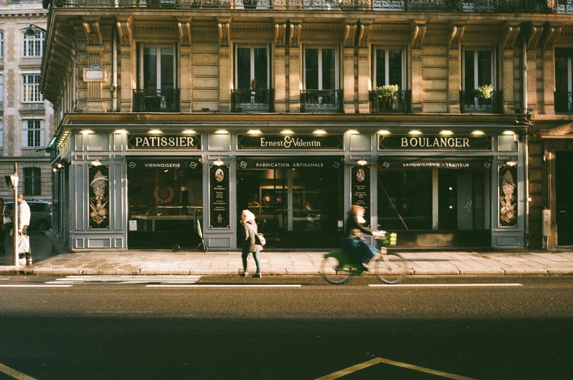Straßenansicht einer Pariser Boulangerie und Patisserie mit Menschen, die an ihrer klassischen Pariser Fassade vorbeigehen und Fahrrad fahren.