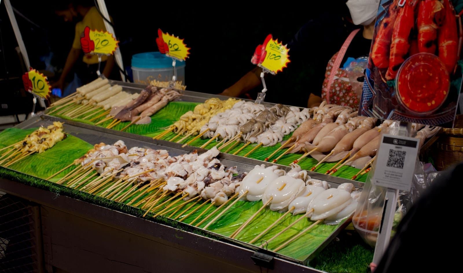 Espetinhos de frutos do mar coloridos expostos numa barraca de comida de rua à noite em Pattaya, com preços e vendedores visíveis ao fundo.