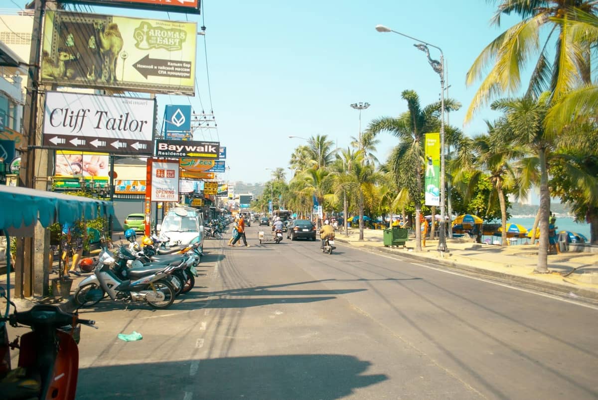 A lively street in Pattaya lined with restaurants, local businesses, food signs, palm trees, and people walking near the beachfront.