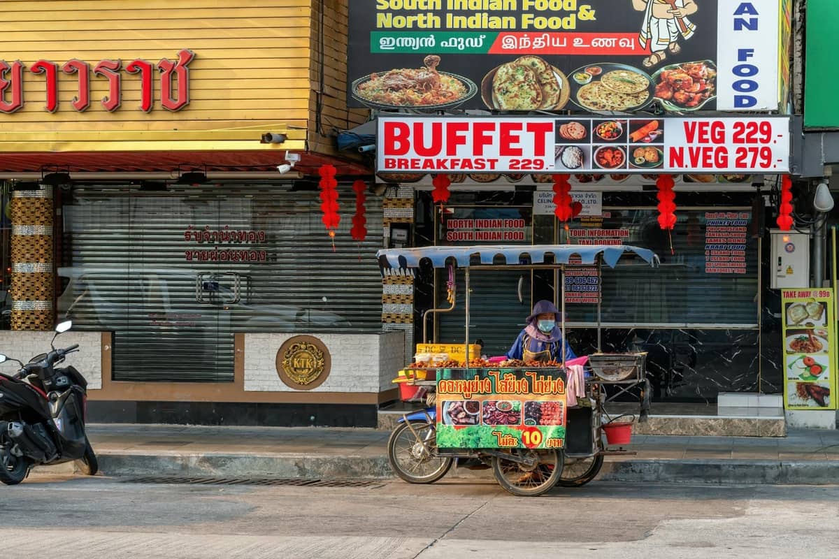 A street food vendor cart in front of restaurants with colorful signs, including South Indian food, reflecting Pattaya's diverse dining scene.