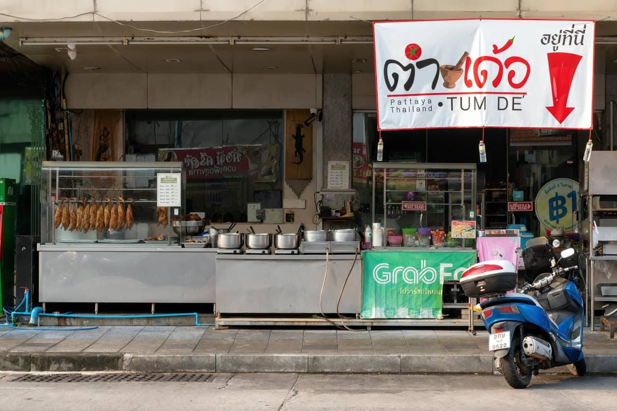Street view of a typical Pattaya shophouse-style Thai restaurant, metal food carts, pots, hanging meats, scooter, and Thai signage.