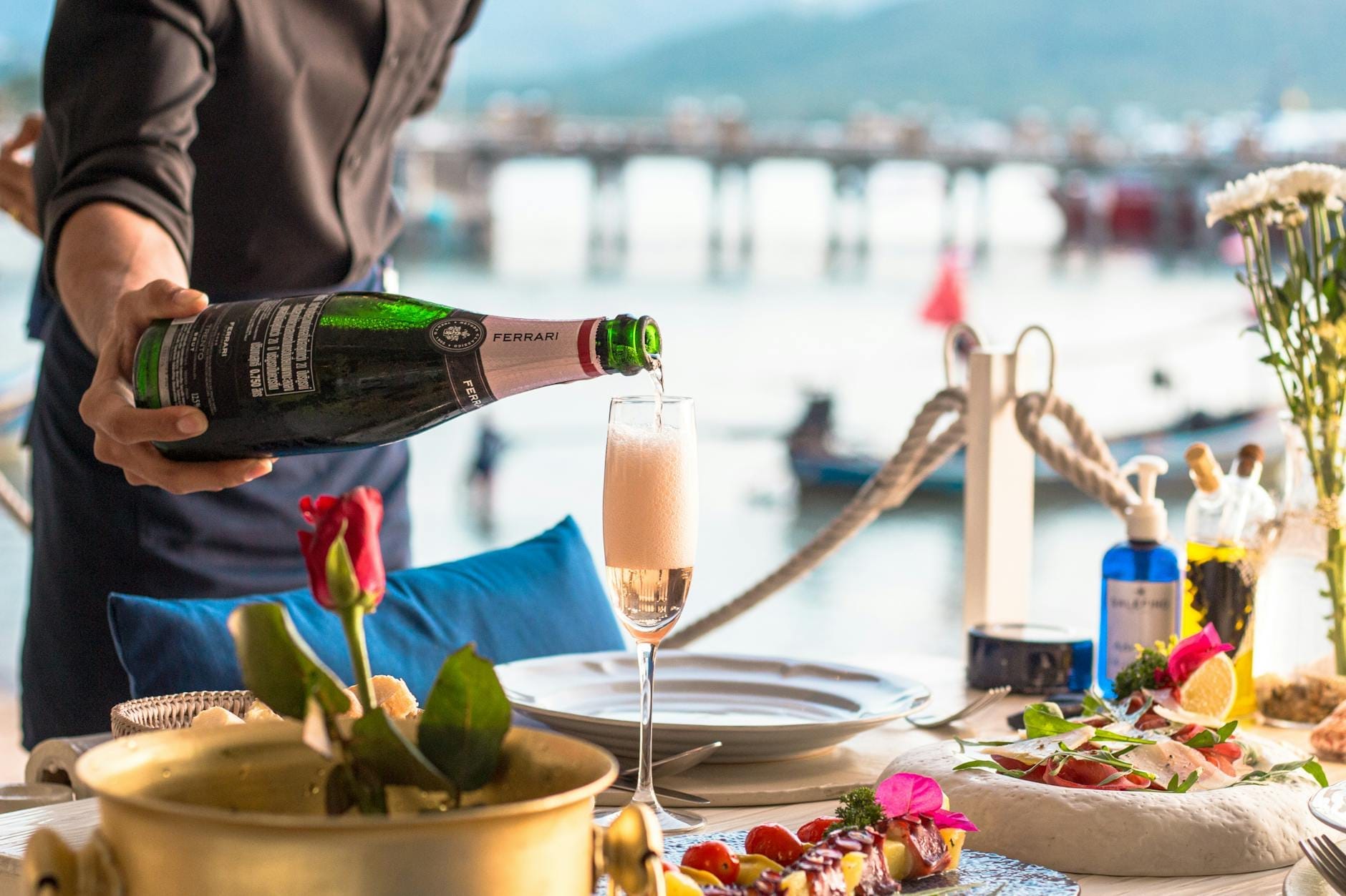 Elegant seaside dining table with fine dishes and champagne being poured, overlooking a pier and water in Phuket.
