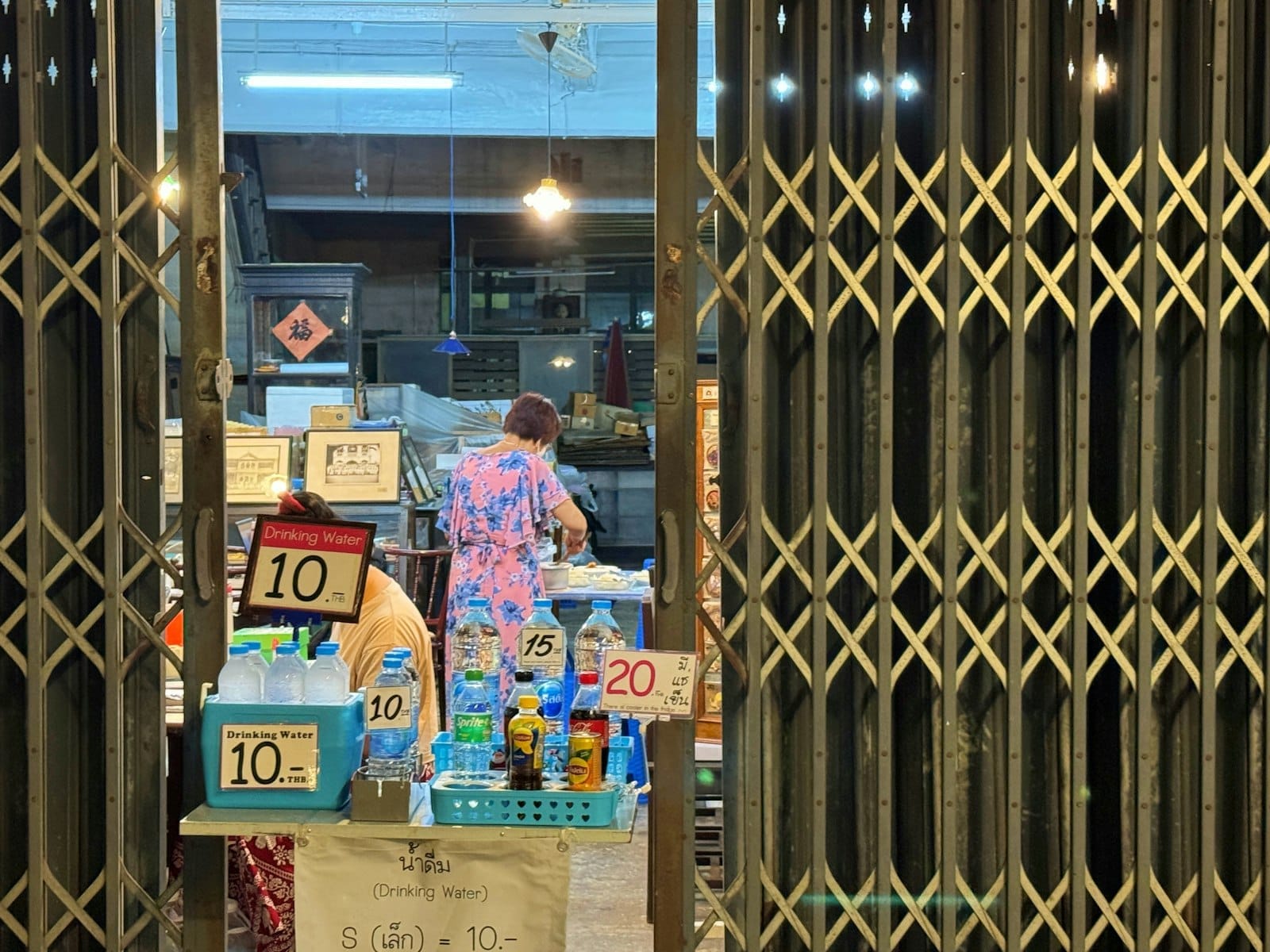 Indoor view of a small food stall in Phuket with bottled drinks priced at 10 to 20 THB, a woman serving food, and a local atmosphere.