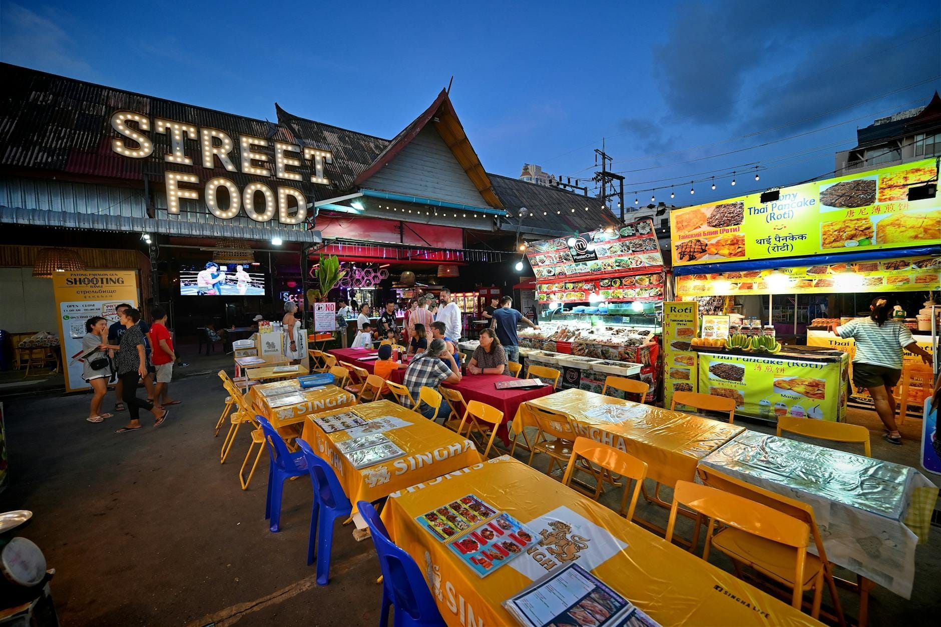 Outdoor street food market in Phuket at dusk with tables, colorful chairs, food stalls, and a large 'Street Food' sign.