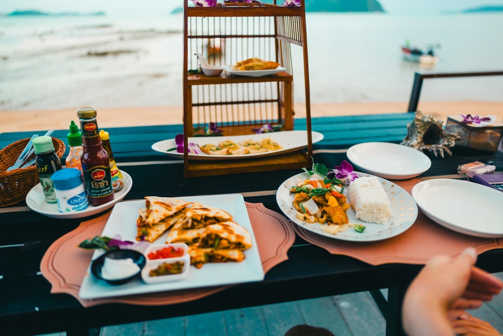 A table at a beachfront restaurant in Phuket set with plates of Thai food, sauces, and drinks with a scenic ocean view.