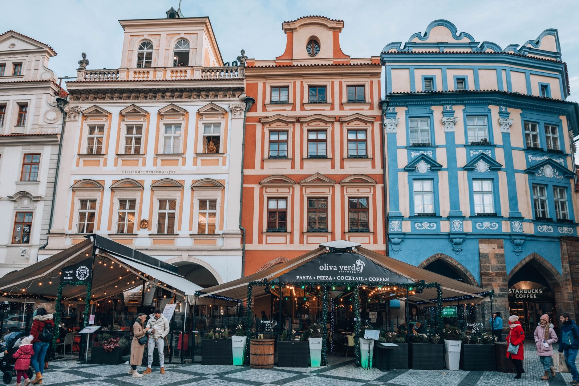 Bunte historische Gebäude mit belebten Straßencafés und Restaurants auf einem lebhaften Platz in Prag, mit Menschen, die durch die Gastronomiebereiche schlendern und dort sitzen.