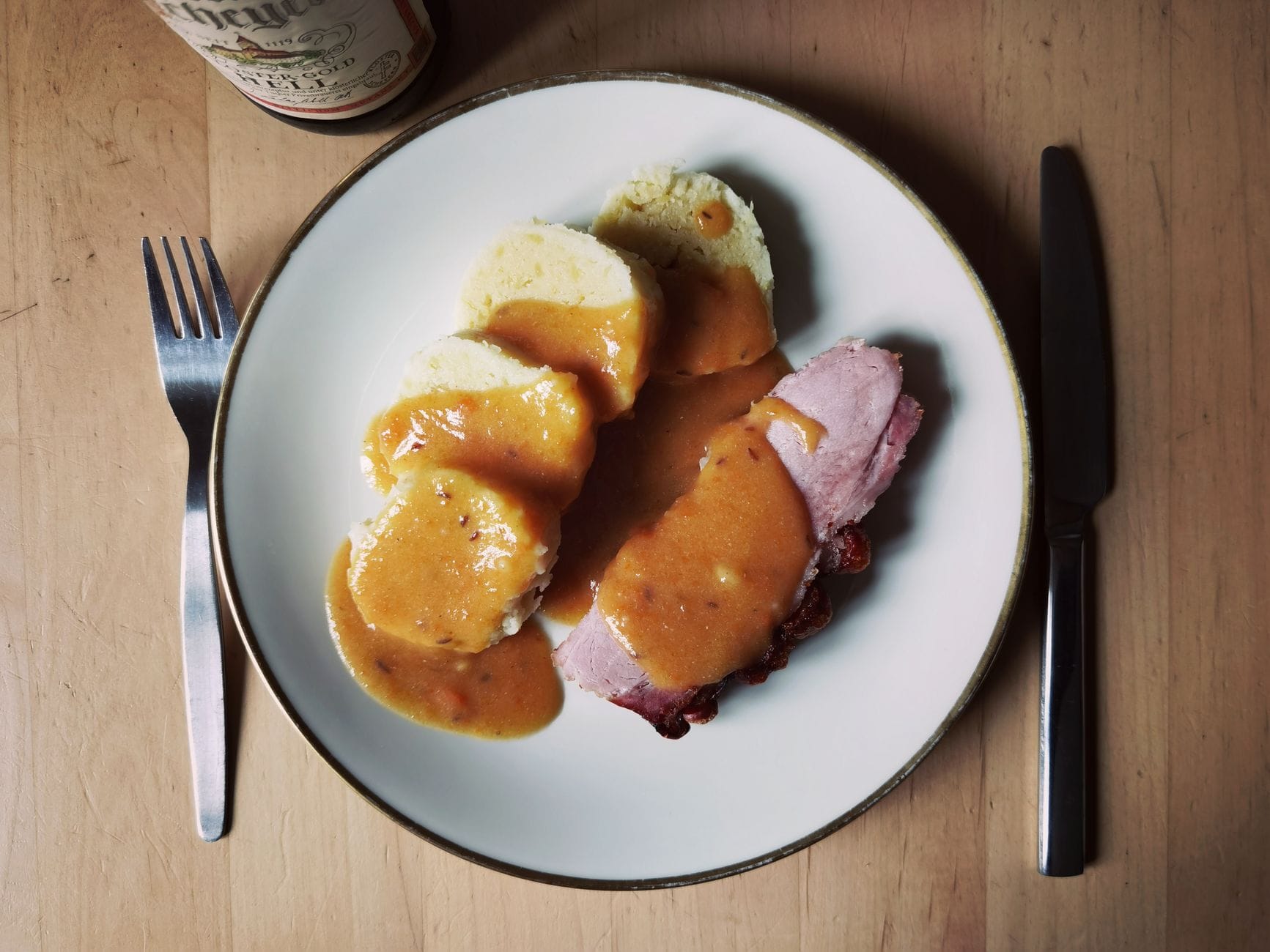 Ein Teller mit geschnittenen Knödeln, Bratenfleisch und reichhaltiger Soße, serviert mit Gabel und Messer, traditionelles tschechisches Essen auf einem Holztisch.