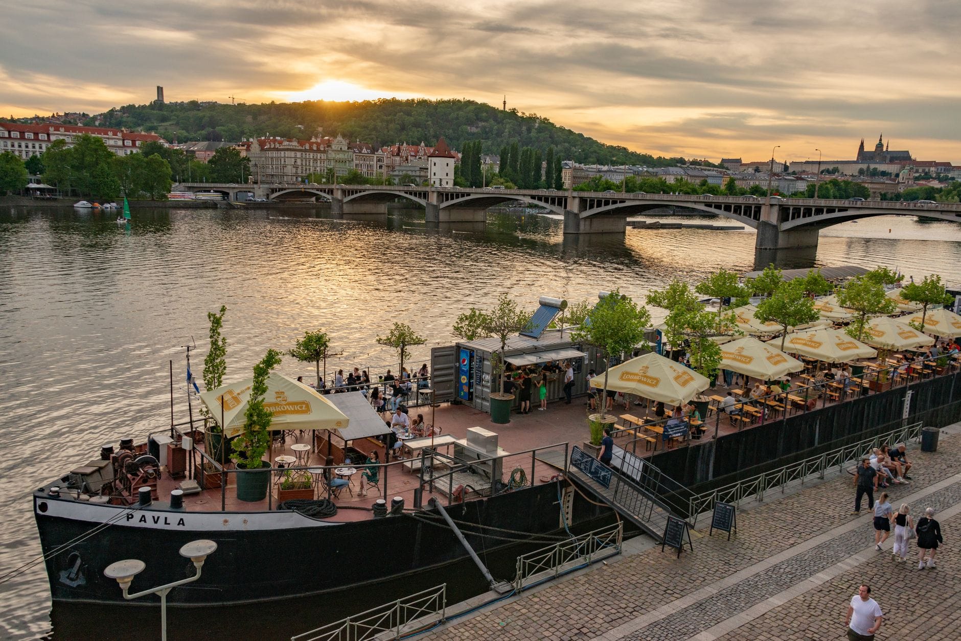 Schwimmendes Restaurant an der Moldau bei Sonnenuntergang, Tische mit Sonnenschirmen, Menschen beim Essen, Stadtbrücke im Hintergrund – ein einladender Anblick für Liebhaber guten Essens.