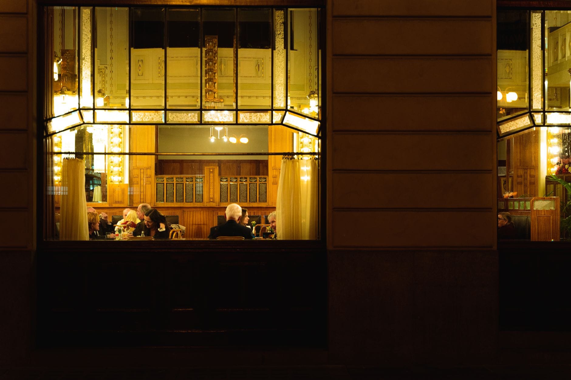 Warmbeleuchteter Blick durch ein großes Restaurantfenster auf Gäste, die eine Mahlzeit in einem stilvollen, klassischen Prager Restaurantinterieur genießen.