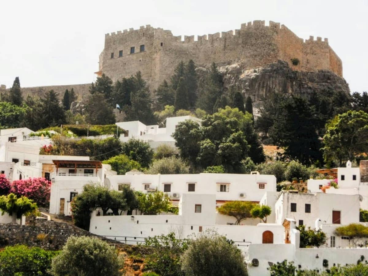 The Acropolis of Lindos rises above whitewashed houses and lush trees in the village of Lindos, Rhodes, under a bright sky.