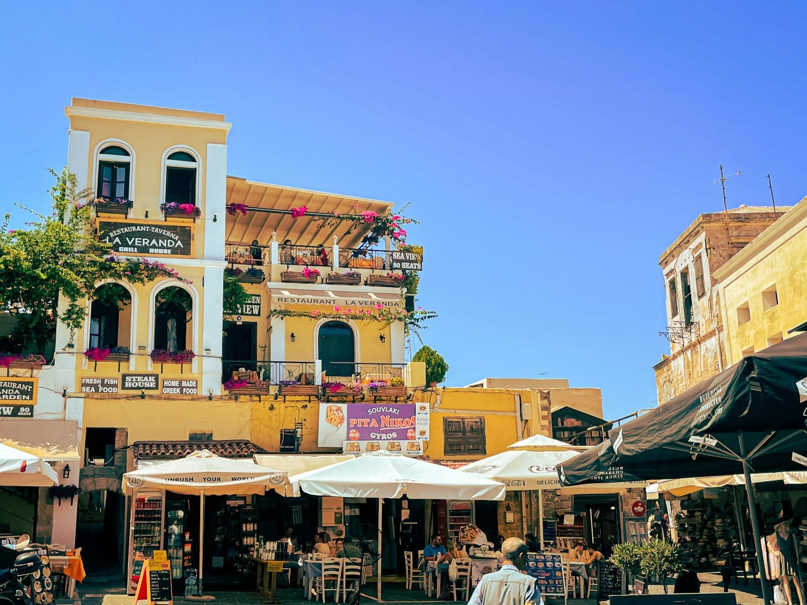 Restaurantes e cafés ao ar livre com guarda-sóis coloridos e edifícios mediterrâneos históricos em uma praça movimentada da Cidade Velha de Rodes sob um céu azul.