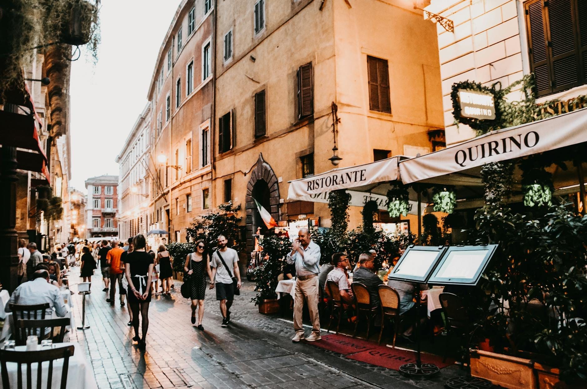 Busy Roman street with people dining outdoors at a ristorante, people walking, and historic buildings in the background at dusk.