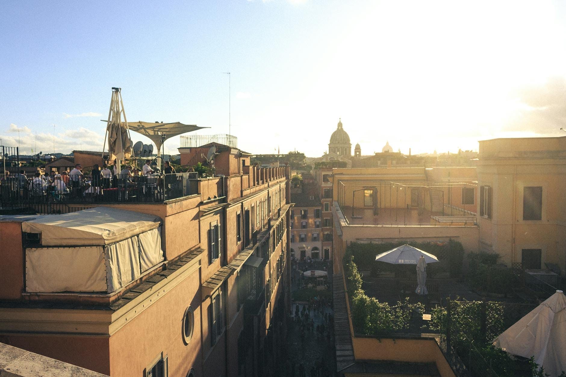 Rooftop restaurant in Rome at sunset with diners enjoying food, historic city buildings, and dome in the background under a bright sky.